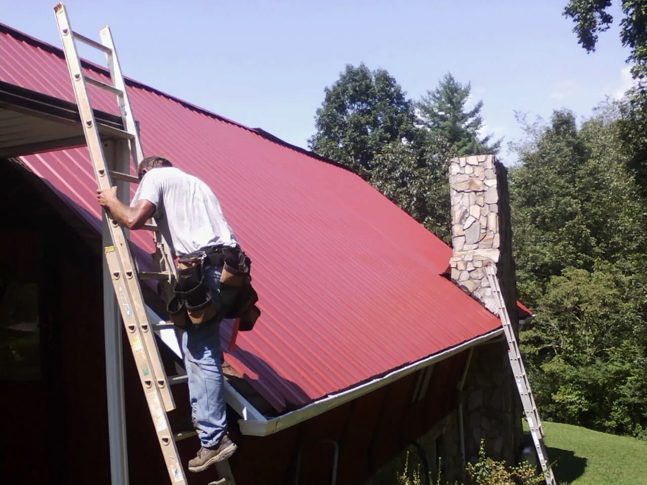 Roofer on a ladder beside a red metal roof and stone chimney — Soffit & Fascia Repair by Roofing Brothers in Lilliwaup